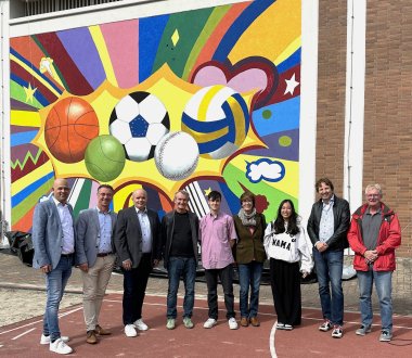 Gruppenbild vor bemalter Rückwand einer Sporthalle für das Europa Gymnasium Wörth. Landrat, Beigeordnete, VG-Bürgermeister, Künstlerinnen und Künstler.