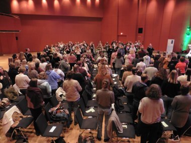 Das Foto zeigt eine Gruppe von Menschen, die von ihren Stühlen aufgestanden sind. Meist mit dem Rücken zum Betrachtenden.