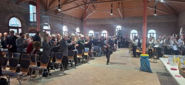 In einer Halle mit Holzdecke läuft ein Mann in der Mitte des Flures nach vorne; die Gäste rechts und links sind von ihren Sitzplätzen (an Tischen) aufgestanden und applaudieren.