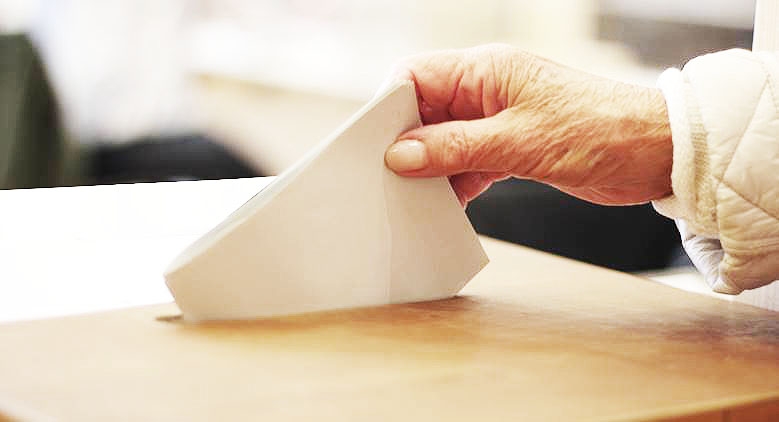 Ein Wahlzettel wird mit der Hand einer älteren Dame in die Wahlurne gesteckt.